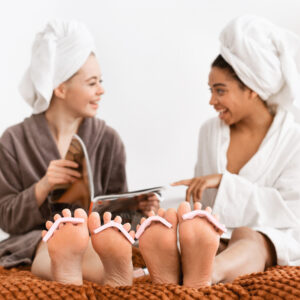 Multiethnic girlfriends in bathrobes sitting on bed and making pedicure at home, reading magazine and talking
